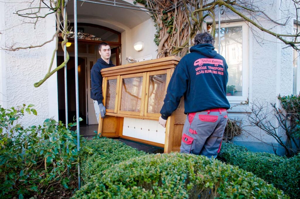 Zwei Männer tragen einen braunen Holzschrank durch einen Eingang, umgeben von grünen Pflanzen.