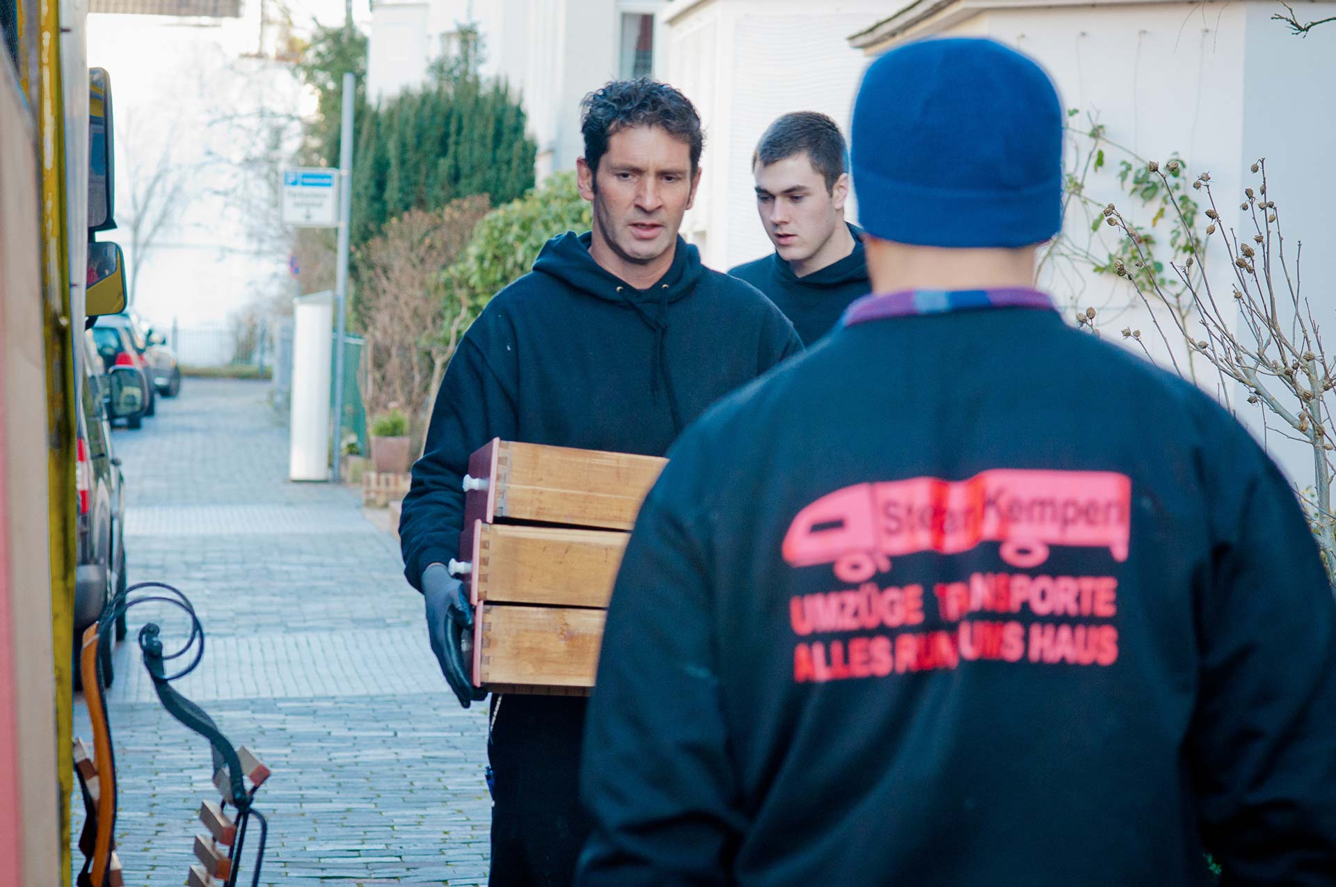 Männer beim Umzug, tragen Holzkisten draußen, konzentrierter Ausdruck, Winterkleidung in Schwarz.