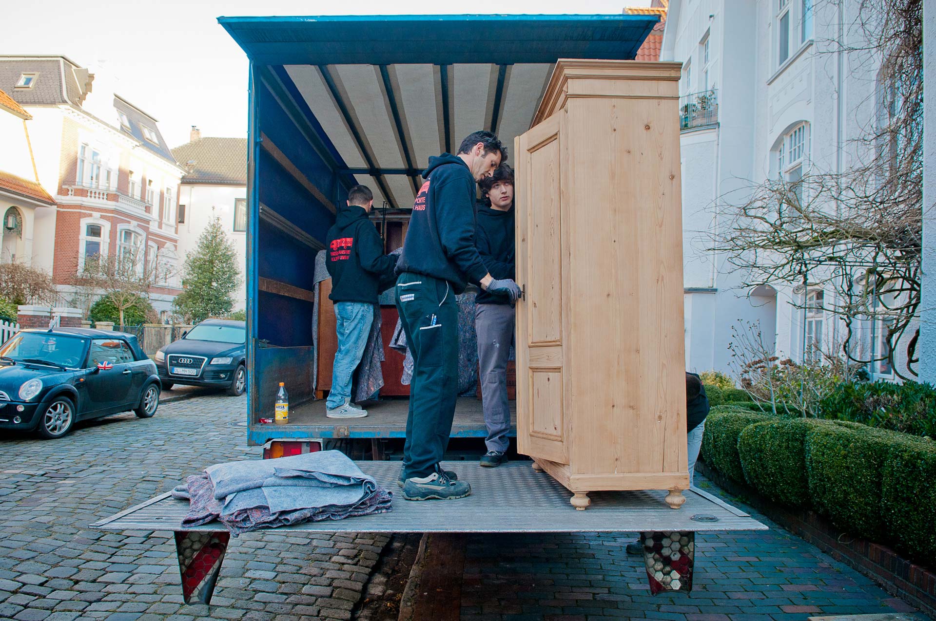 Personen bewegen einen Holzschrank in einen LKW. Sie wirken konzentriert und tragen dunkelblaue Kleidung.