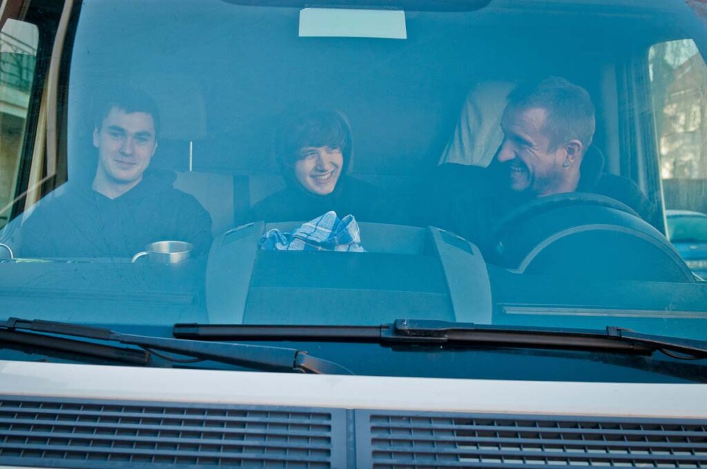 Drei Personen lächeln in einem Auto, blauer Himmel draußen, Becher und Tuch auf dem Armaturenbrett.
