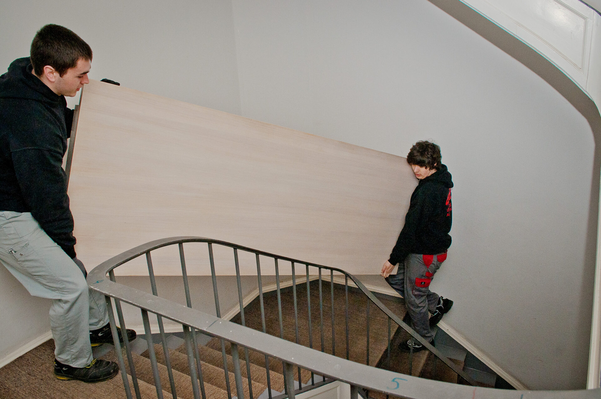Zwei Personen tragen eine große Holztür eine Treppe hinauf, beide konzentriert und in dunkler Kleidung.