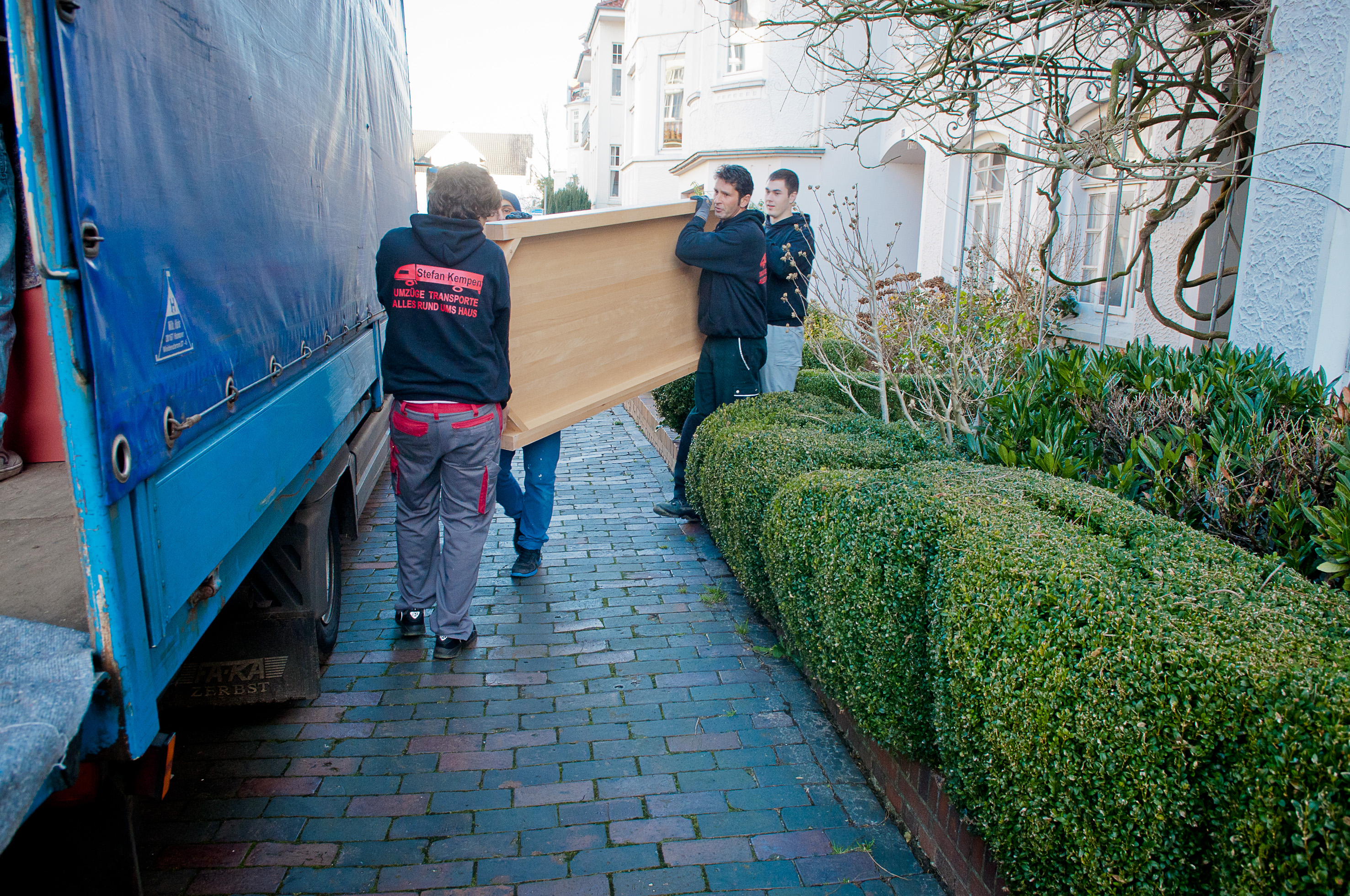 Vier Personen tragen ein Möbelstück in einen LKW. Es ist tagsüber, die Straße ist gepflastert.